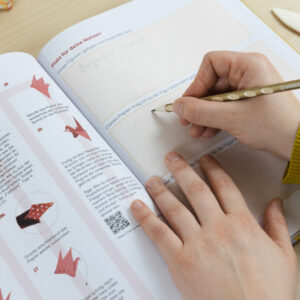 Geöffnetes Buch. Es zeigt links eine Seite in der die Faltschritte gezeigt werden. Auf der rechten Seite sind Platz für Notizen. Auf dieser Seite werden gerade mit einem Bleistift erste Gedanken geschrieben.
