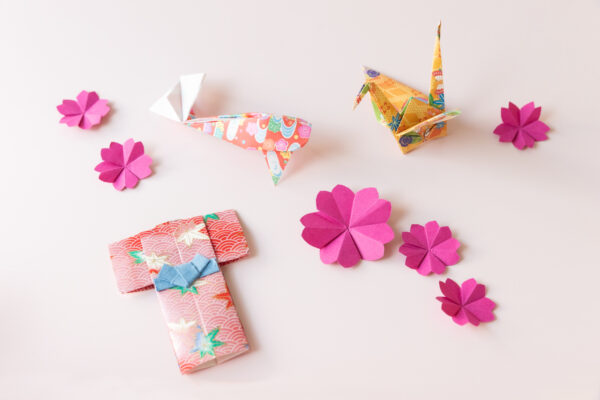 Auf einem weißen Tisch liegen verschiedene Origami-Figuren: 6 Kirschblüten, ein Kranich, ein Koi und ein Kimono.