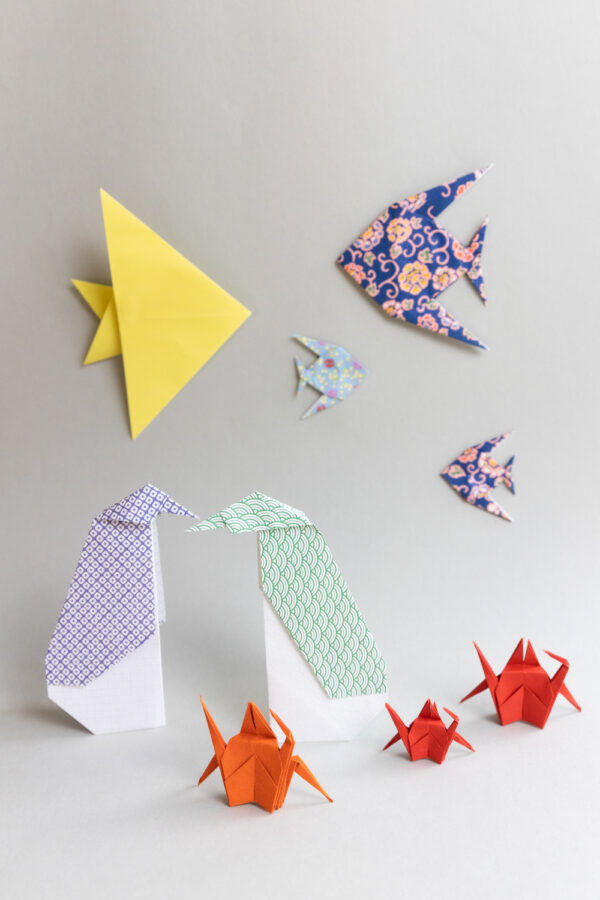 Auf einem weißen Tisch stehen verschiedene Origami Figuren: Zwei Pinguine und drei Krebse. An der Wand im Hintergrund kleben 4 Origami Fische.