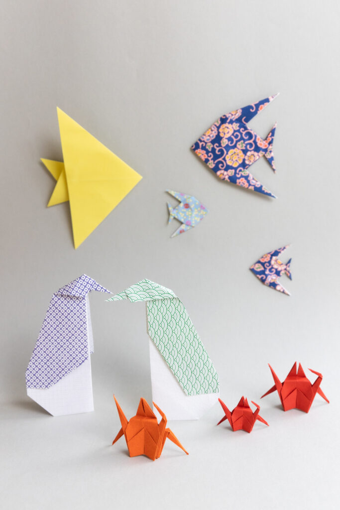 Auf einem weißen Tisch stehen verschiedene Origami Figuren: Zwei Pinguine und drei Krebse. An der Wand im Hintergrund kleben 4 Origami Fische.
