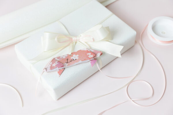 Auf einem hellen Tisch liegt ein Geschenk, eingewickelt in weißem Geschenkpapier und einer cremefarbenen Schleife umwickelt. Um das Geschenk liegt noch weiteres Geschenkpapier und -band.