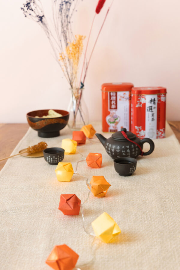 Auf einem Tisch steht chinesisches Teegeschirr, eine Schüssel mit Knabbereien, und im Hintergrund zwei Teedosen und Trockenblumen. Im Fokus liegt eine Lichterkette die mit bunten Origami Bällen verziert ist.