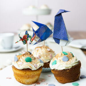 Ein Tisch wurde mit weißem Kaffee-Geschirr gedeckt. Im Fokus steht ein Teller und darauf sind drei Muffins mit Creme und Streuseln. In die Muffins wurden mit Holzstäbchen Origami Figuren gesteckt: Ein Krebs, ein Pinguin und ein Fisch.