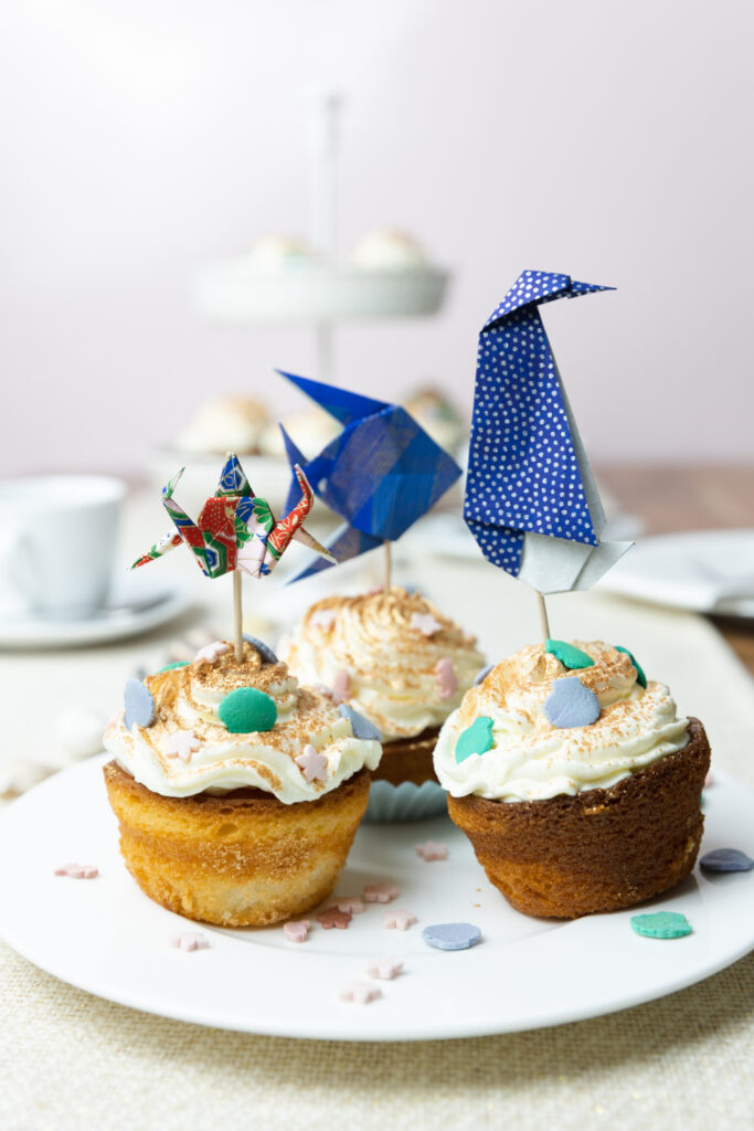 Ein Tisch wurde mit weißem Kaffee-Geschirr gedeckt. Im Fokus steht ein Teller und darauf sind drei Muffins mit Creme und Streuseln. In die Muffins wurden mit Holzstäbchen Origami Figuren gesteckt: Ein Krebs, ein Pinguin und ein Fisch.