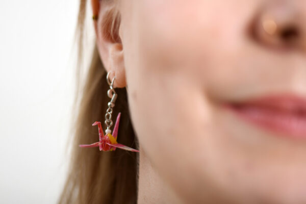 Pinker Origami Kranich Ohrringe aus Papier hängt an einem Ohr einer blonden Frau.