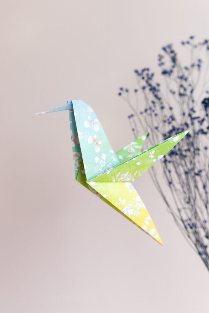 Origami Kolibri fliegt durch das Bild. Im Hintergrund sind Trockenblumen.
