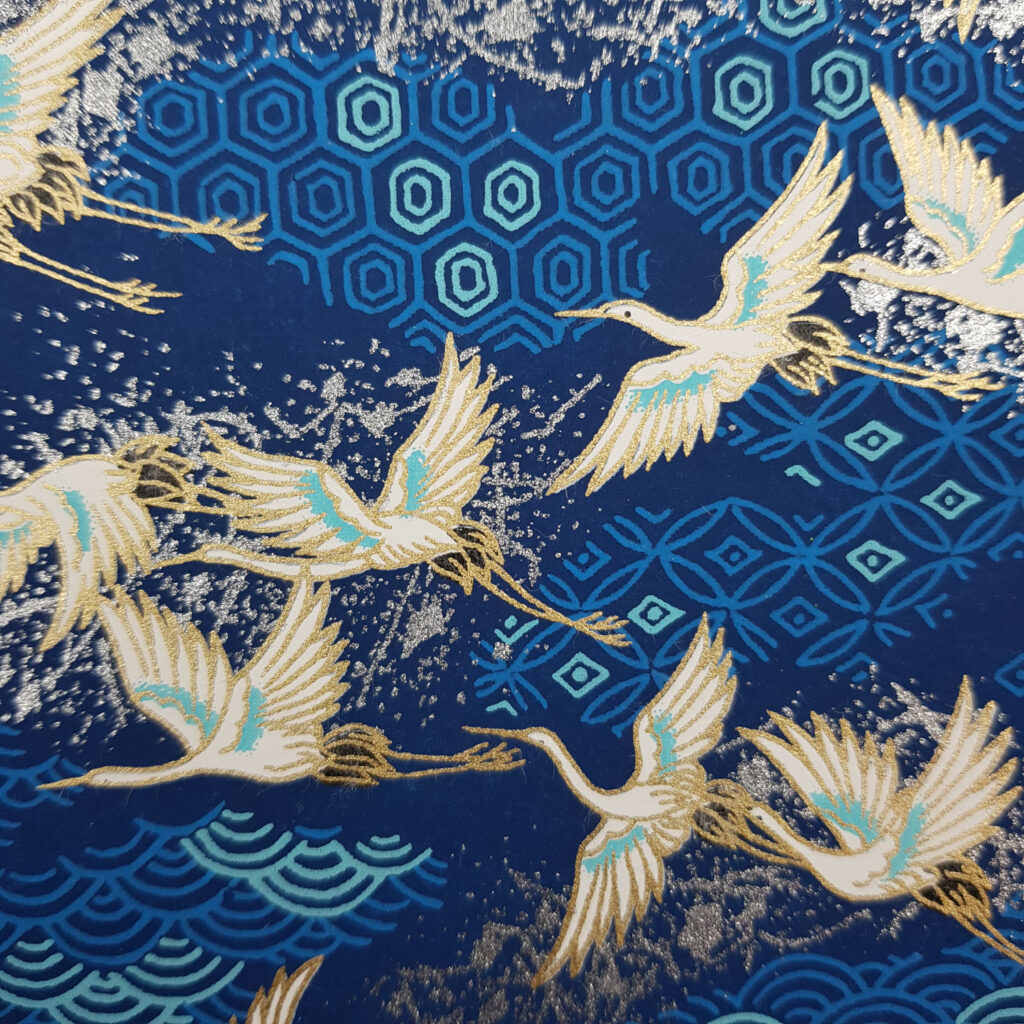 Blaues Origami Papier mit weißen Kranichen die fliegen und Gold umrandet sind.